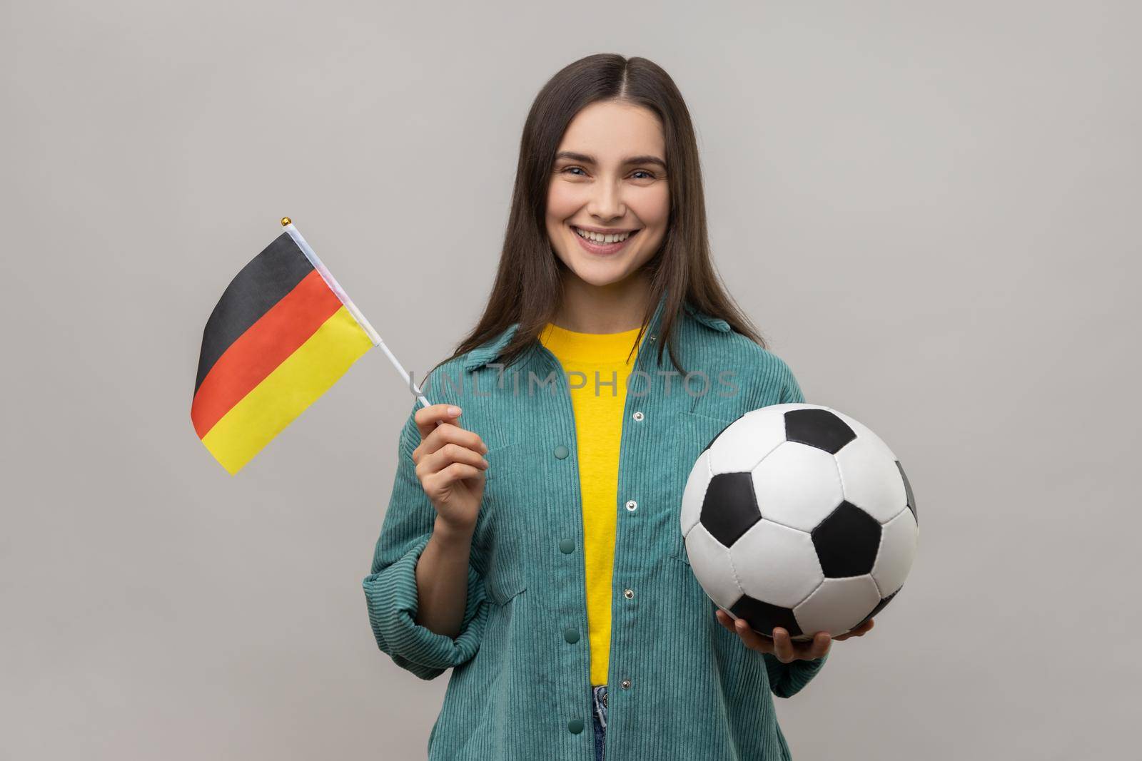 Smiling satisfied woman holding in hands flag of Germany and football ball, supporting favourite team on championship, wearing casual style jacket. Indoor studio shot isolated on gray background.