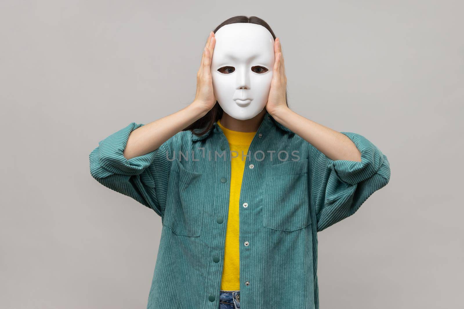 Portrait of unknown woman holding white mask, standing covering face, multiple personality disorder, wearing casual style jacket. Indoor studio shot isolated on gray background.