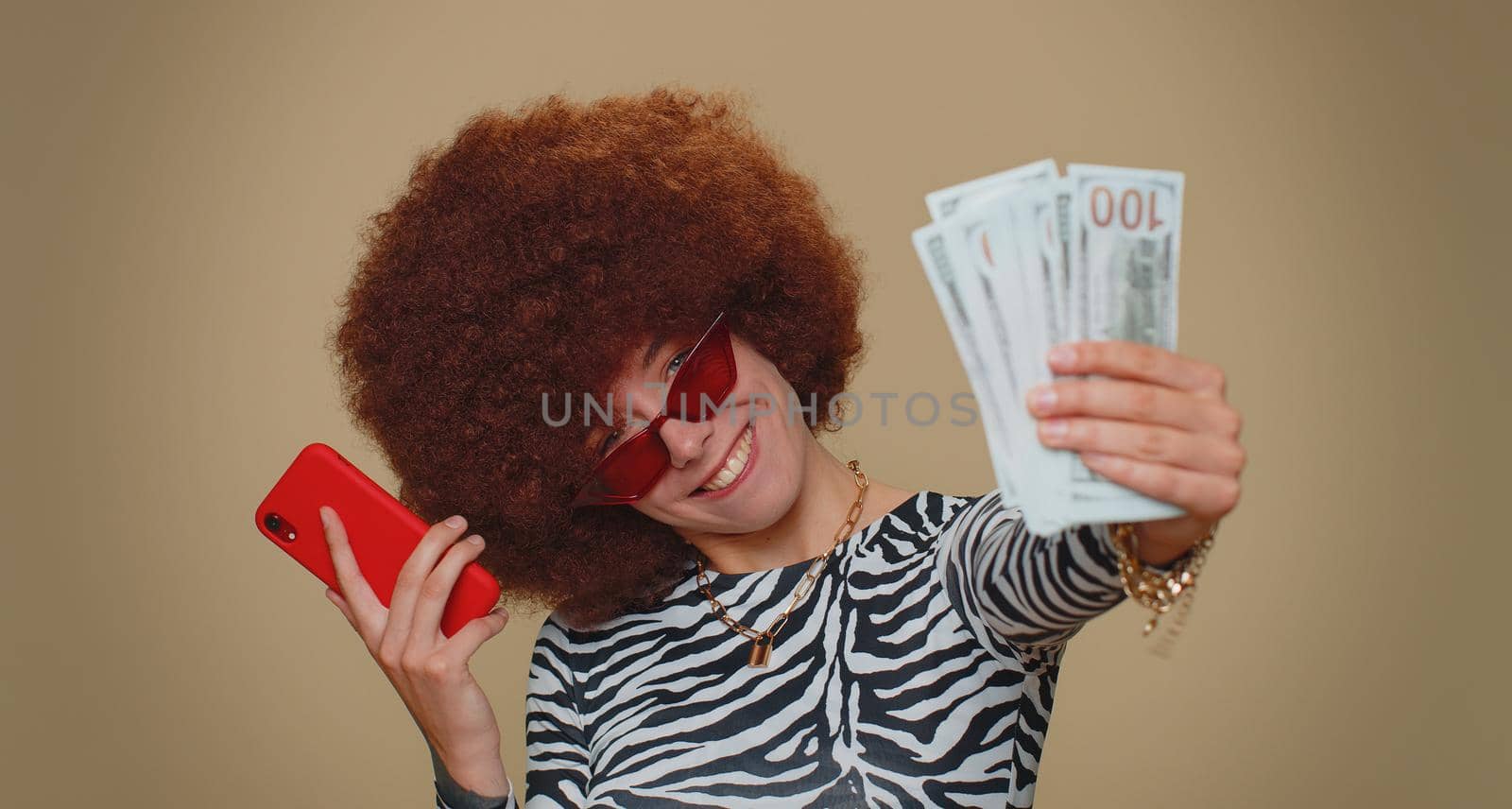 Happy young woman with lush wig looking smartphone display sincerely rejoicing win, receiving money dollar cash banknotes, success lottery luck. Adult stylish female girl on beige studio background