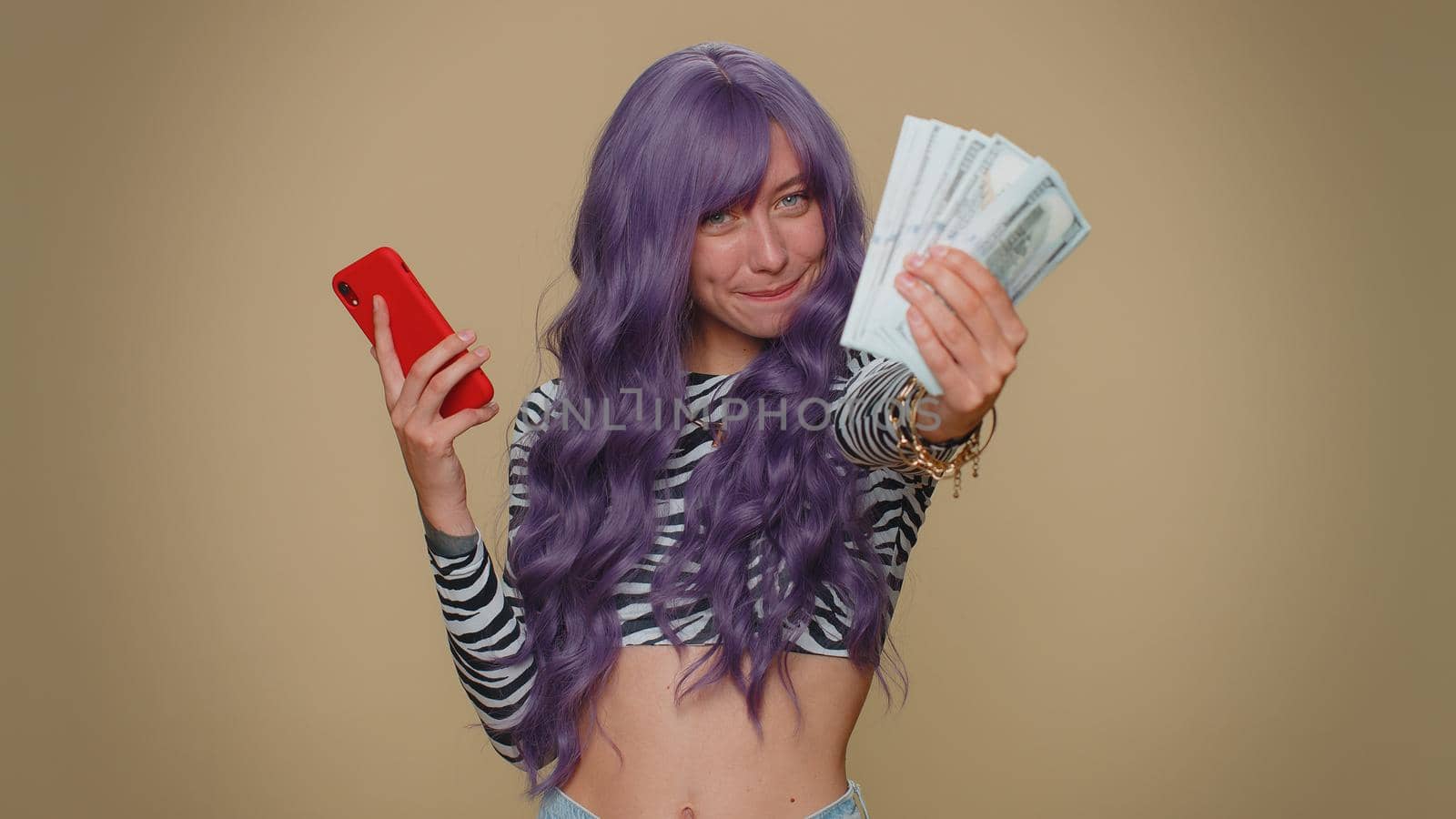 Happy young woman with lush wig looking smartphone display sincerely rejoicing win, receiving money dollar cash banknotes, success lottery luck. Adult stylish female girl on beige studio background