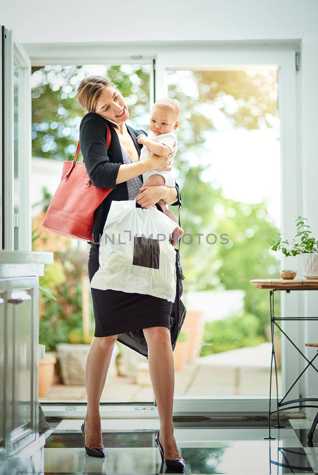 The reality of being a working Mom. a busy businesswoman carrying a shopping bag and her baby while talking on the phone on her return from work. by YuriArcurs