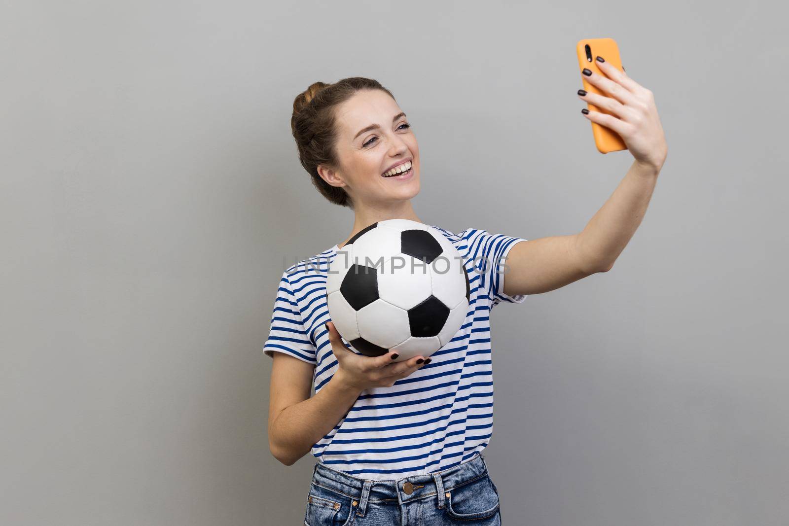 Portrait of smiling woman wearing striped T-shirt making selfie, having video call or broadcasting livestream with ball in hand. Indoor studio shot isolated on gray background.