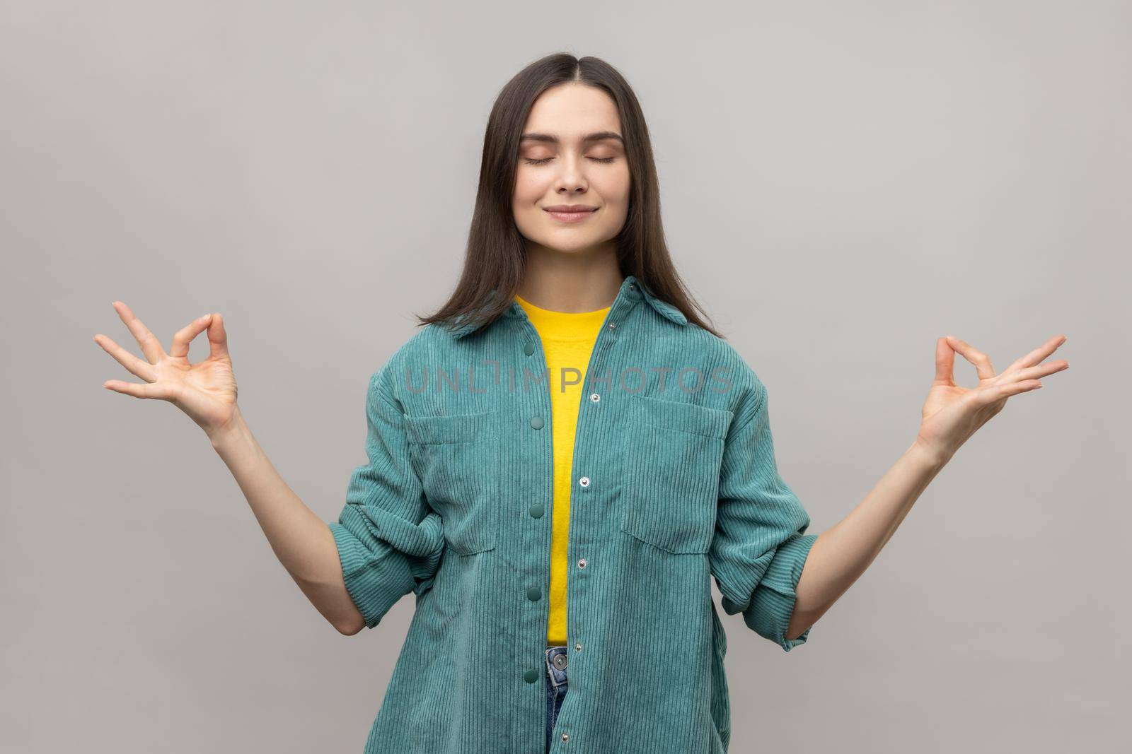 Portrait of peaceful young woman relaxing and doing meditation gesture with fingers, practicing yoga, wearing casual style jacket. Indoor studio shot isolated on gray background.
