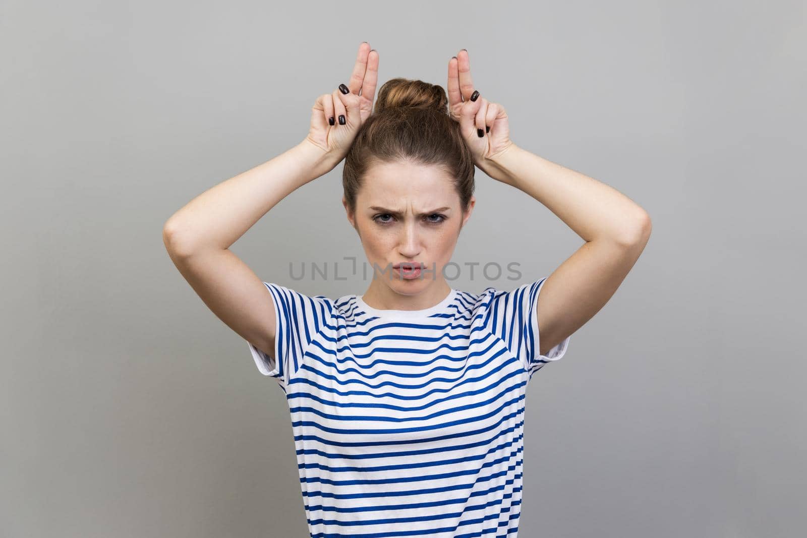 Portrait of angry bully woman wearing T-shirt showing bull horns with fingers on head, antler gesture, threatening with hostile view, ready to attack. Indoor studio shot isolated on gray background.
