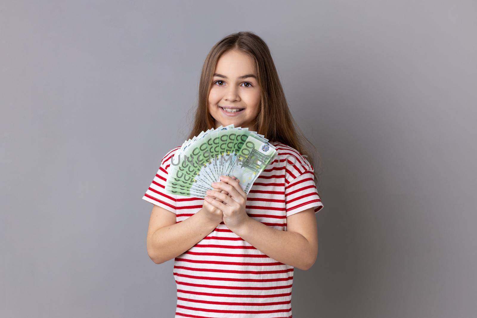 Portrait of satisfied little girl wearing striped T-shirt holding at fan of euro banknotes, showing big money and looking at camera. Indoor studio shot isolated on gray background.