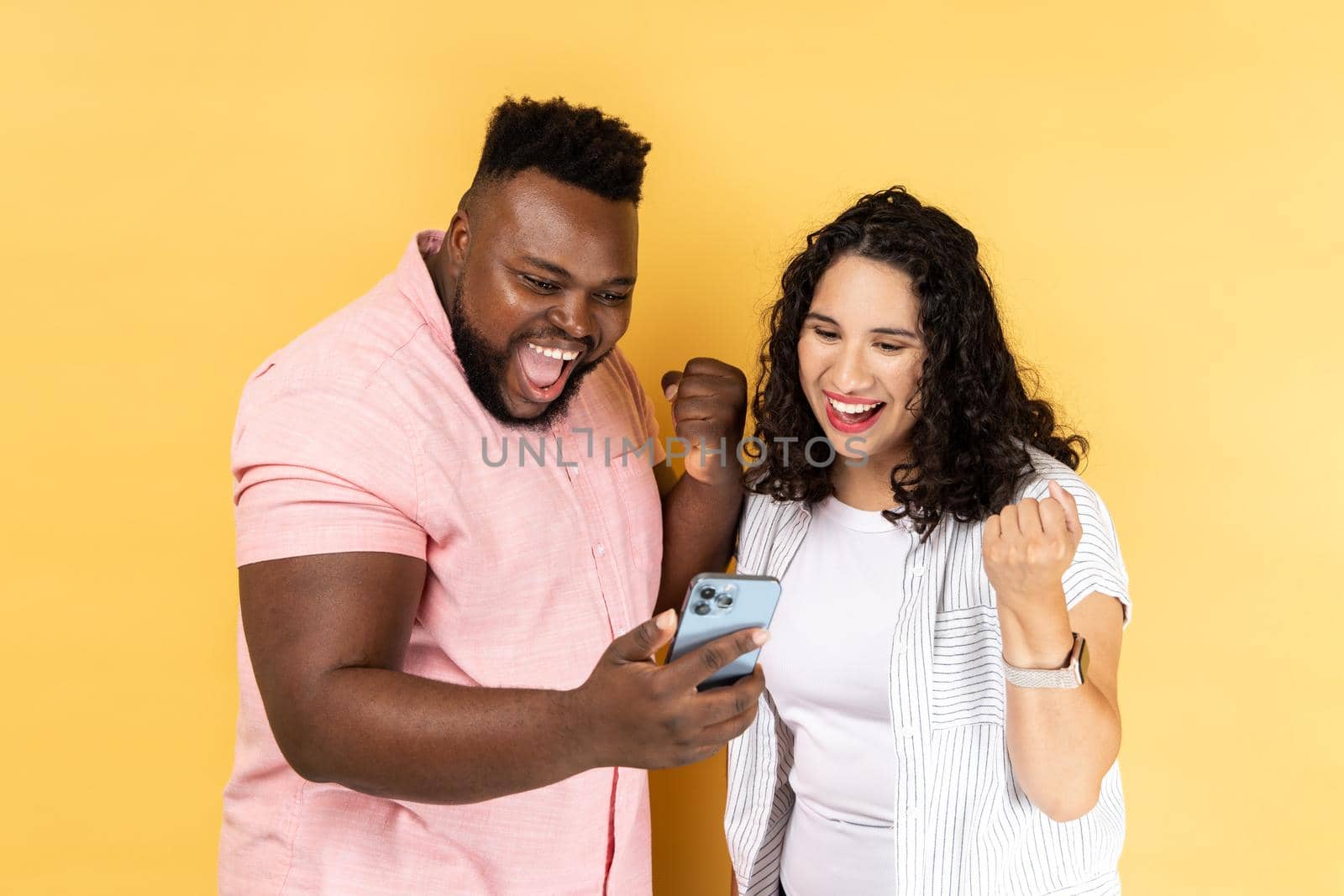 Portrait of excited extremely happy young couple in casual clothing standing together and looking at smart phone, clenched fists, celebrating victory. Indoor studio shot isolated on yellow background