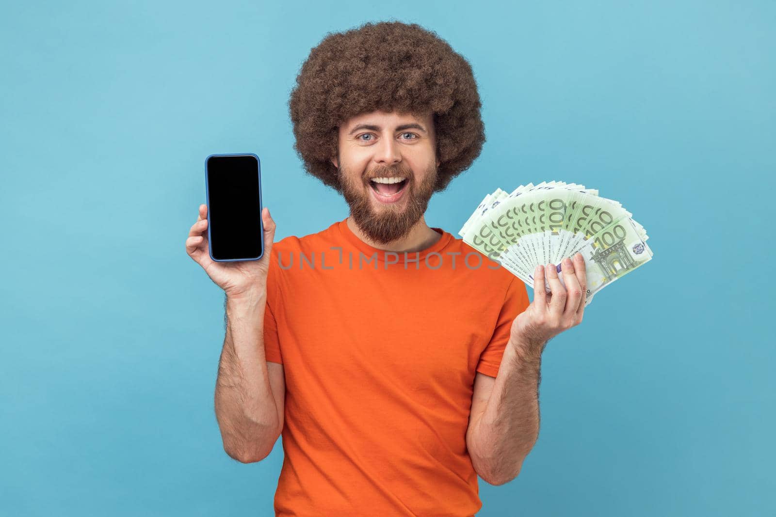 Portrait of satisfied man with Afro hairstyle wearing orange T-shirt showing euro banknotes and smart phone with empty display for promotional text. Indoor studio shot isolated on blue background.