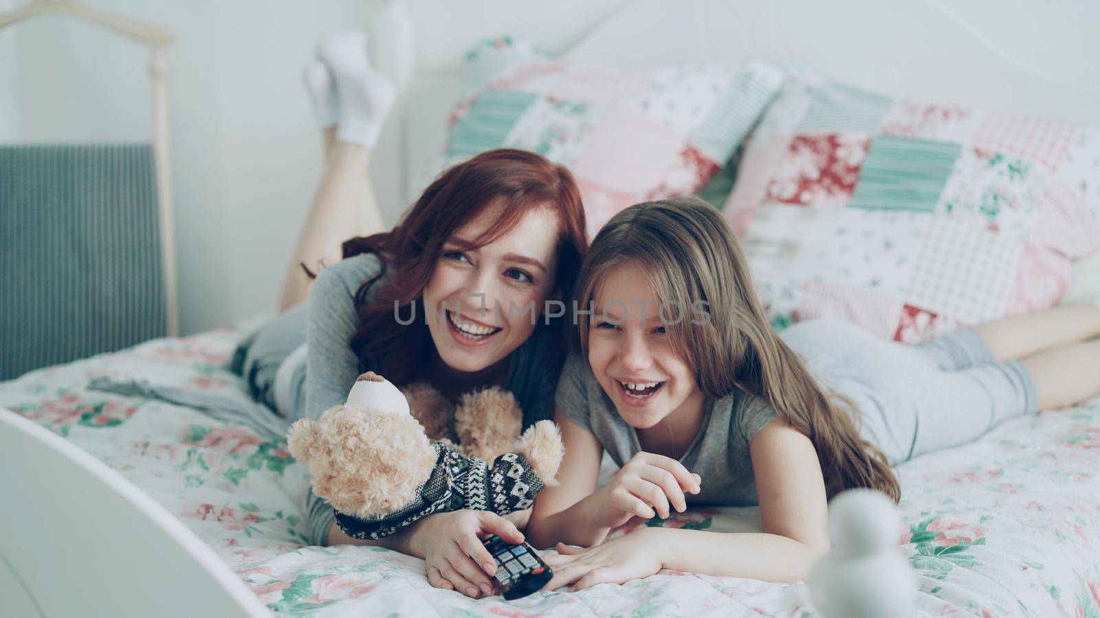 Laughing happy mother and cute daughter watching funny cartoon movie on TV while lying on bed at home in the morning and have fun by silverkblack