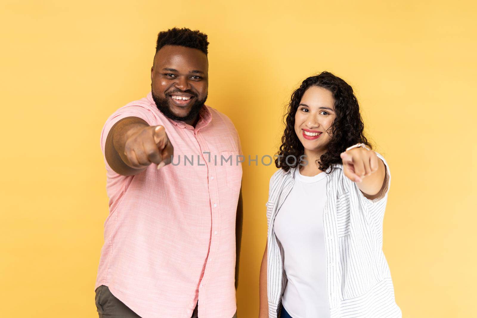 Portrait of smiling satisfied couple in casual clothing standing together and pointing at camera with index fingers, expressing positive emotions. Indoor studio shot isolated on yellow background.