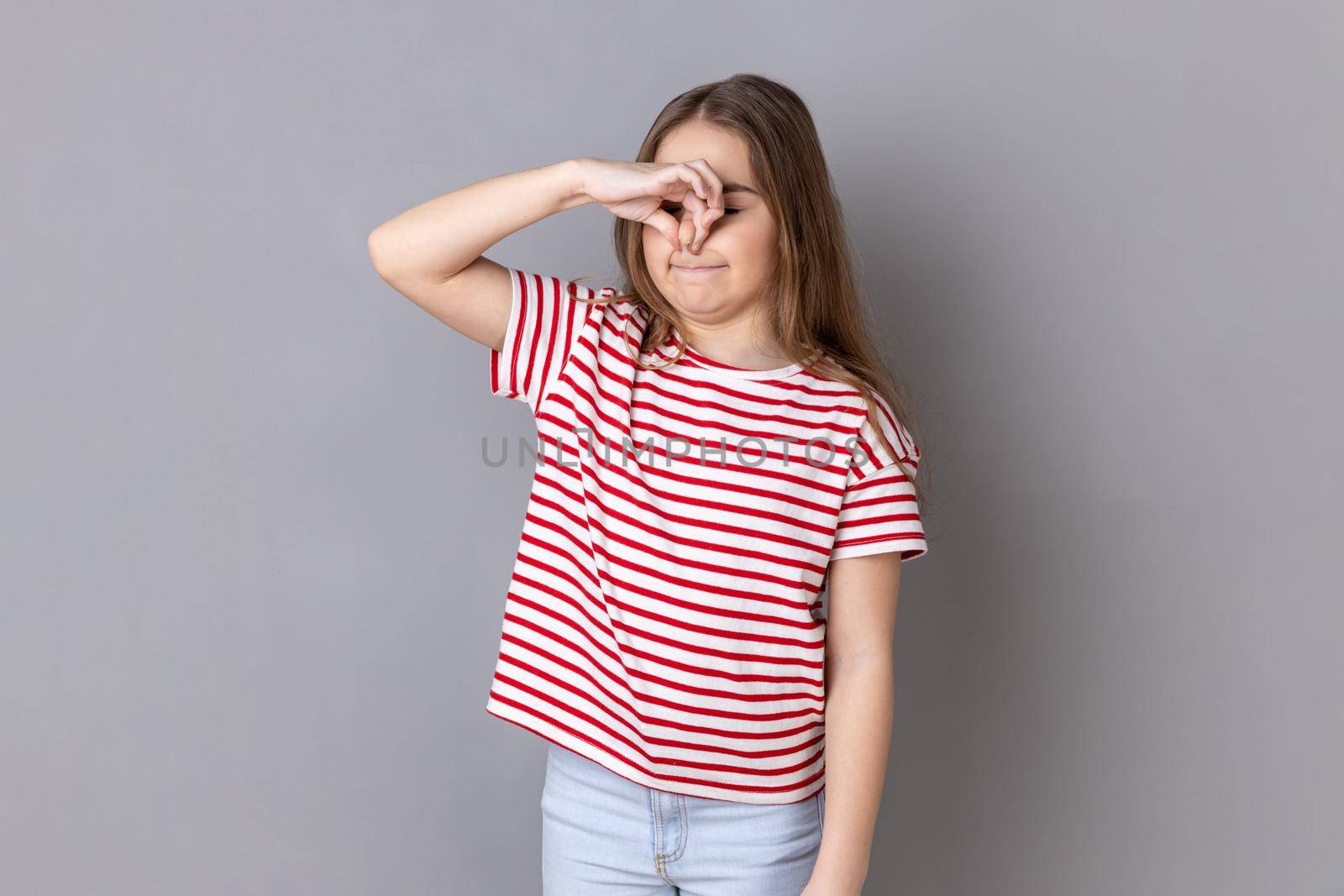 Portrait of confused little girl wearing striped T-shirt pinching her nose to hold breath, feeling repulsion and disgust to unpleasant odor. Indoor studio shot isolated on gray background.