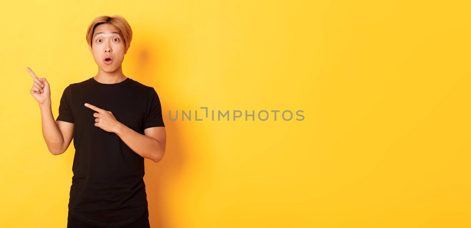 Portrait of impressed and excited handsome asian guy in black t-shirt, react to your logo, pointing fingers upper left corner by Benzoix