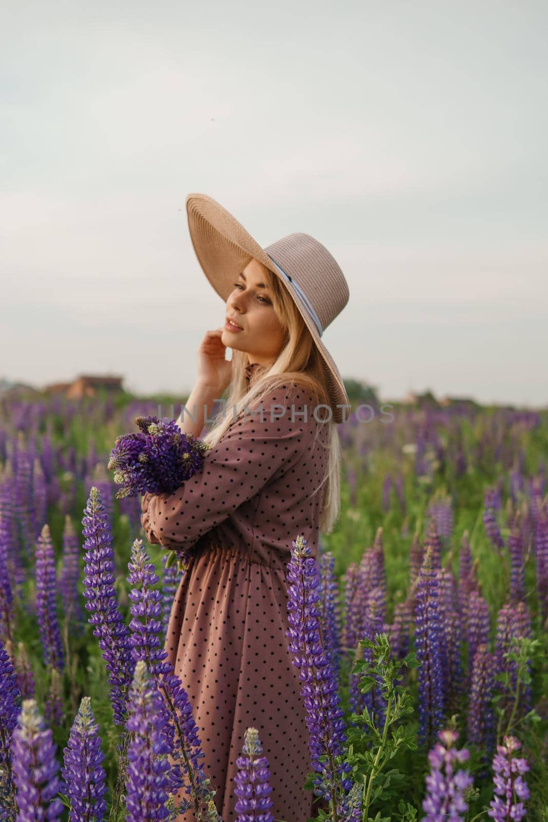 A beautiful woman in a straw hat walks in a field with purple flowers. A walk in nature in the lupin field.