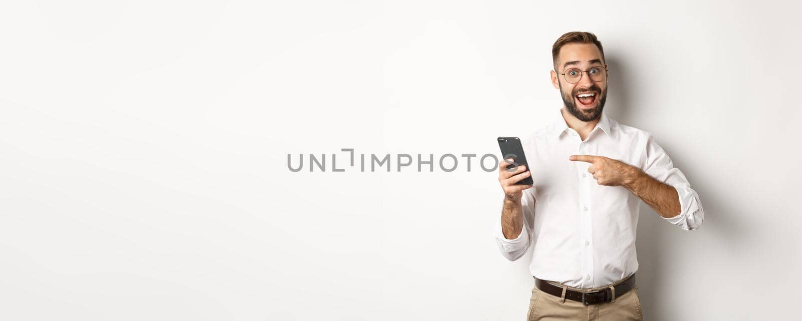 Man looking excited and pointing finger at mobile phone, showing good online offer, standing over white background.