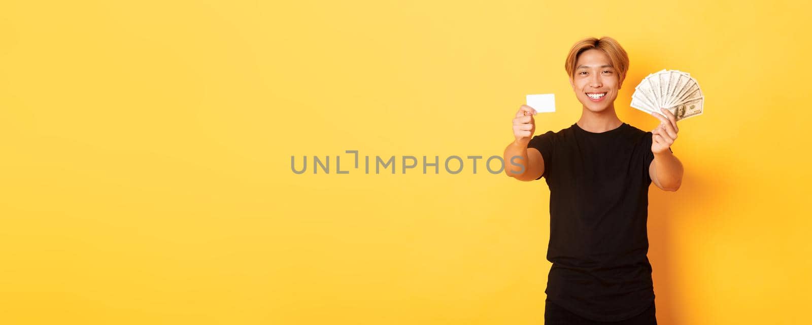 Satisfied attractive korean guy with blond hair, showing money and credit card, smiling happy, yellow background.