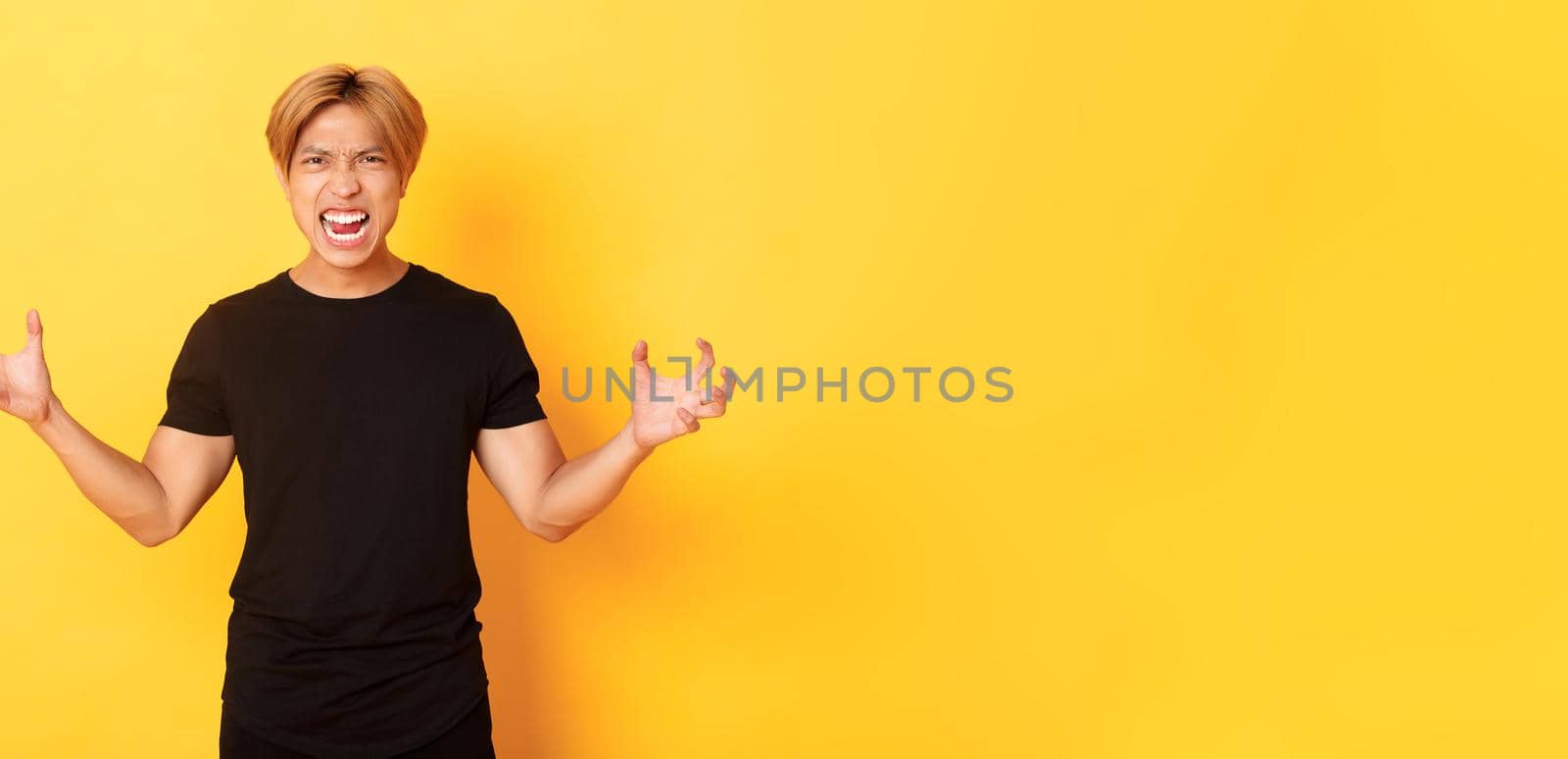 Portrait of outraged, pissed-off asian man shaking hands hateful and grimacing, shouting angry, standing over yellow background.