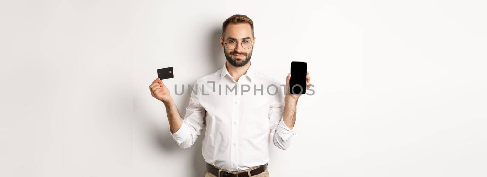 Handsome bearded man showing mobile phone and credit card, shopping online, standing over white background.