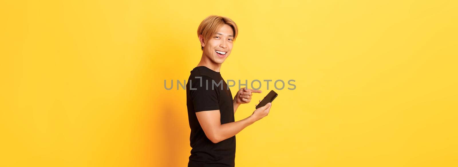 Portrait of pleased smiling handsome asian man, standing in profile and pointing finger at smartphone, recommend app, standing yellow background by Benzoix