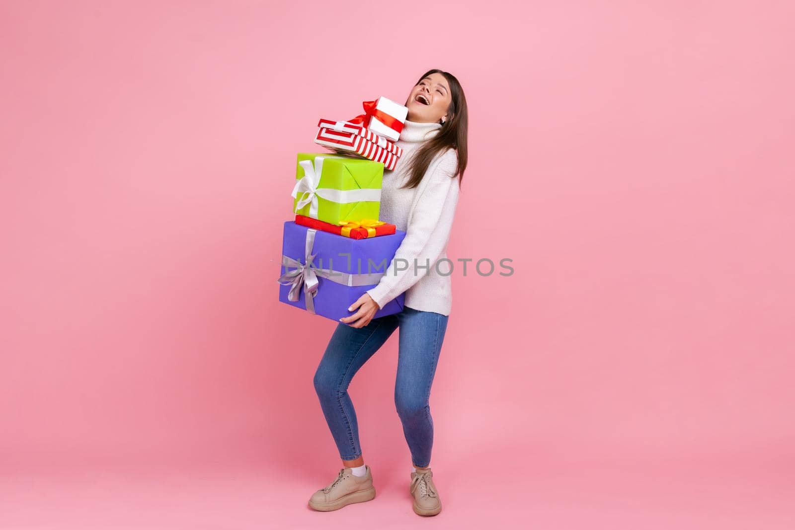 Full length portrait of happy brunette woman holding heavy stack of presents, celebrating holiday, wearing white casual style sweater. Indoor studio shot isolated on pink background.