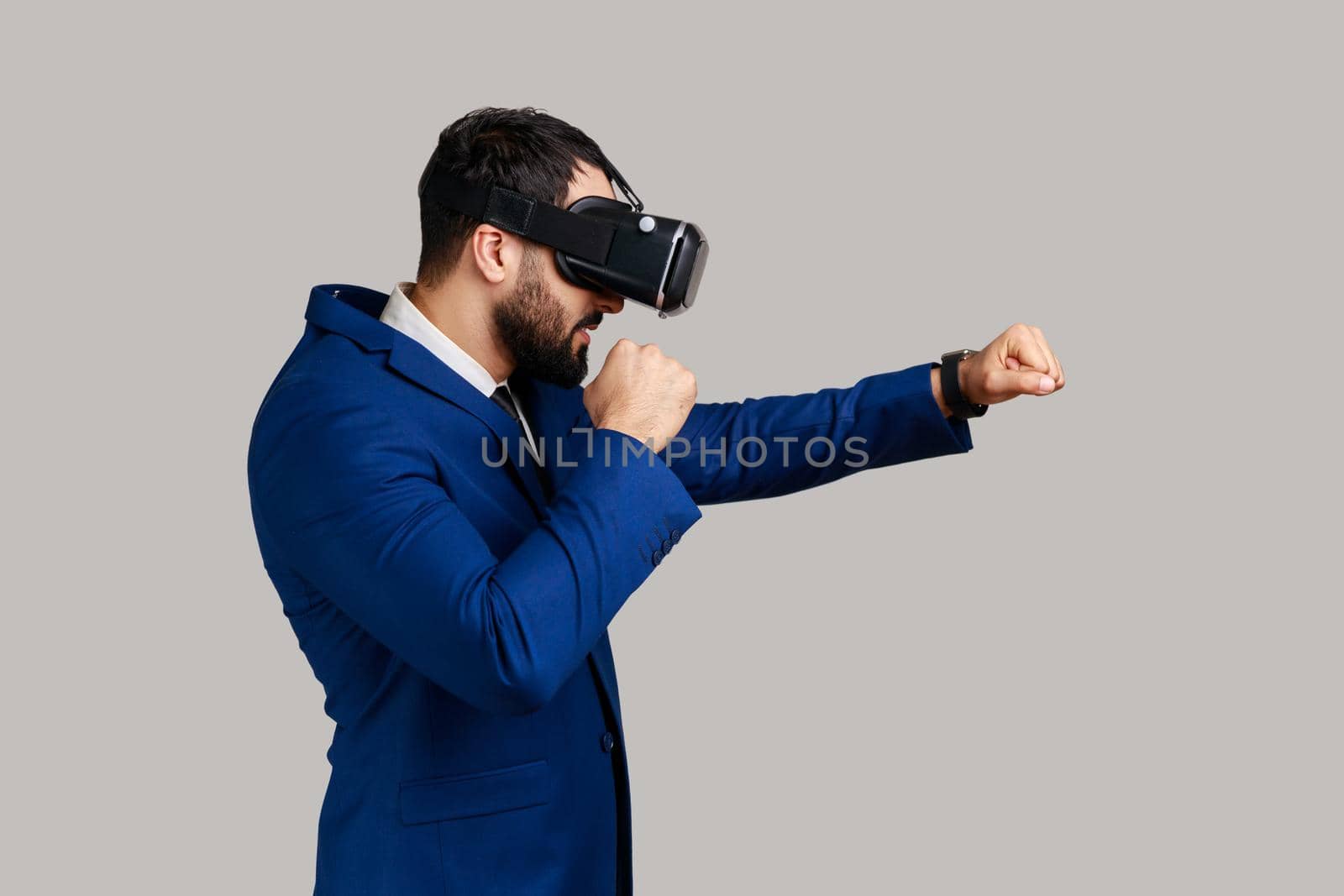 Side view of man virtual reality glasses on head playing fighting game, holding clenched fists up ready to boxing, wearing official style suit. Indoor studio shot isolated on gray background.