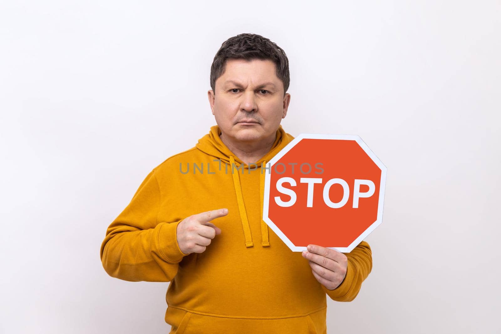 Portrait of middle aged strict bossy man pointing at Stop road traffic sign as symbol of prohibition, wearing urban style hoodie. Indoor studio shot isolated on white background.