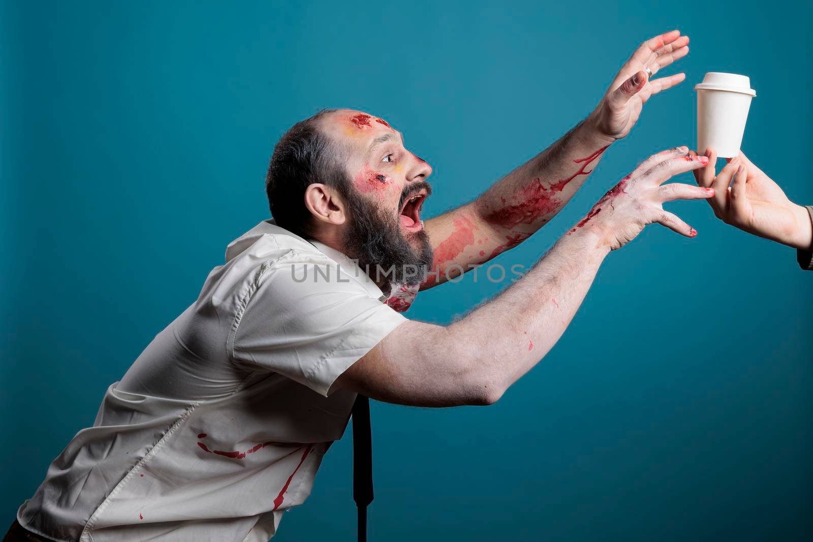 Halloween evil monster wanting coffee cup in studio over blue background, going after desired drink and acting aggressive. Cruel undead zombie with bloody scars wishing for beverage.