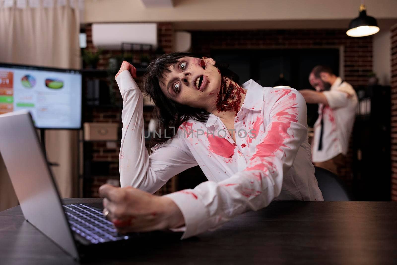 Horror cruel zombie using laptop at desk, undead corpse trying to work on computer in startup office. Creepy aggressive brain eating monster looking terrifying and horrible, sinister danger.