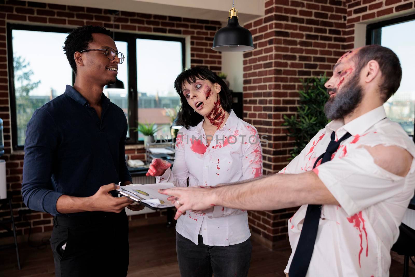 Young adult talking to evil killer zombies in startup office, chatting with brain eating cruel monsters. Man and woman corpses with bloody wounds and scars having conversation with person, macabre.