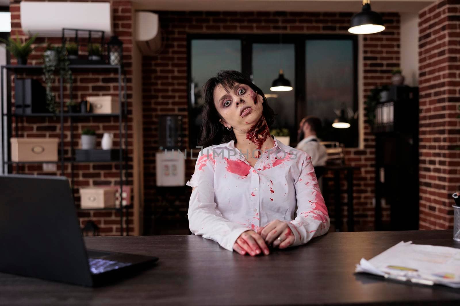 Portrait of creepy woman zombie at office desk, sitting and looking terrifying with wounds and scars. Scary cruel walking dead corpse eating brain and being aggressive, sinister gory nightmare.