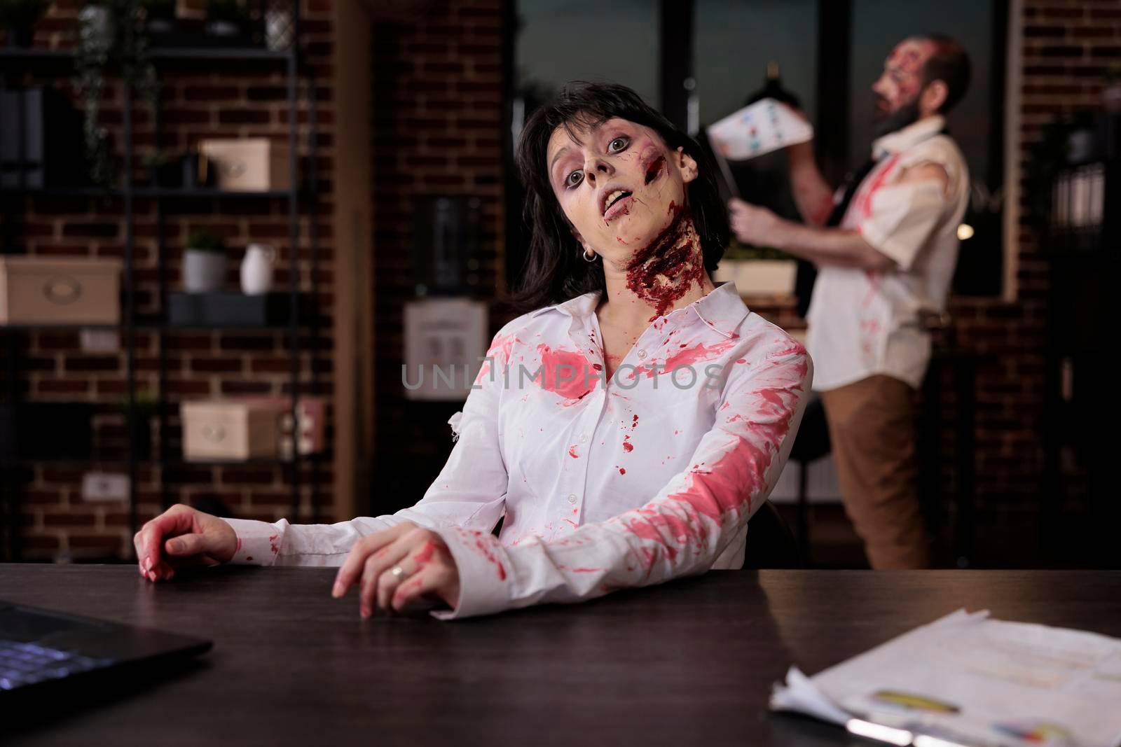 Portrait of scary corpse with wounds in office, sitting at desk to work on laptop being terrifying and spooky. Cruel possessed brain eating monster with bloody mouth open, walking dead devil.