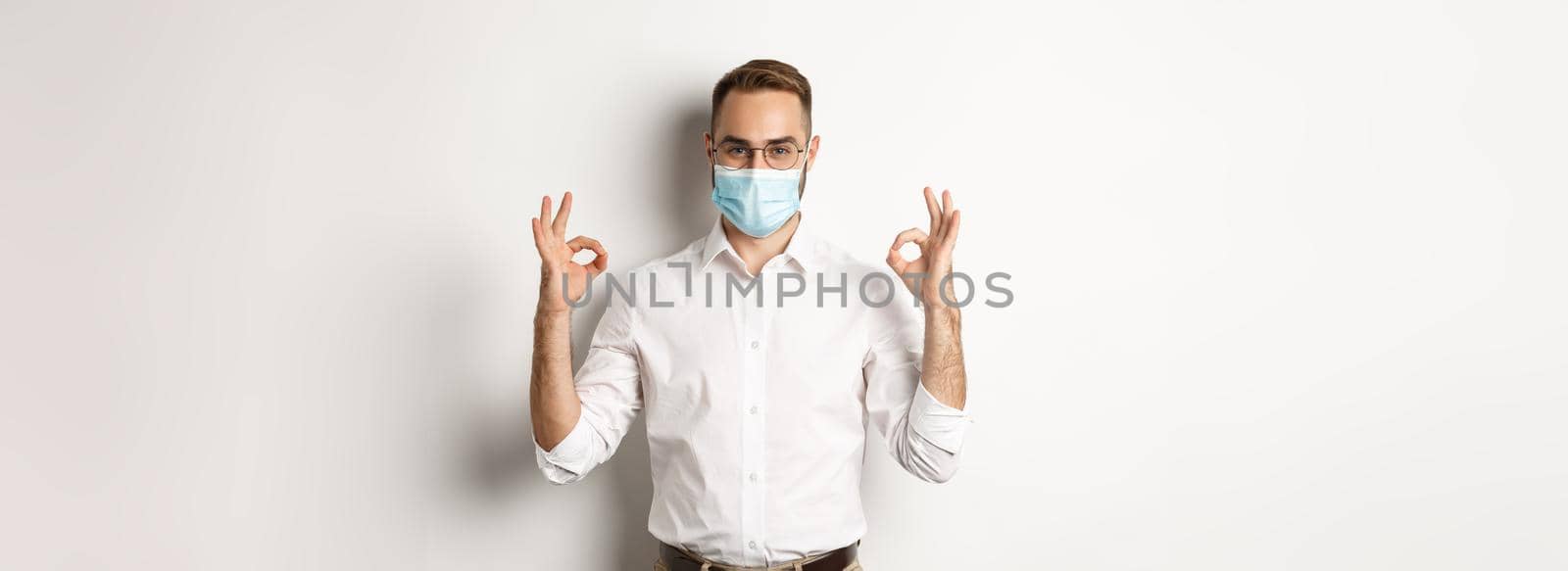 Covid-19, social distancing and quarantine concept. Confident businessman wearing medical mask and showing okay signs in approval, white background by Benzoix