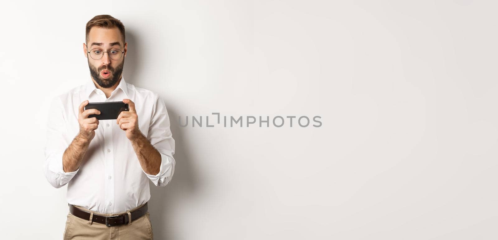 Man looking amazed at mobile phone, standing against white background.