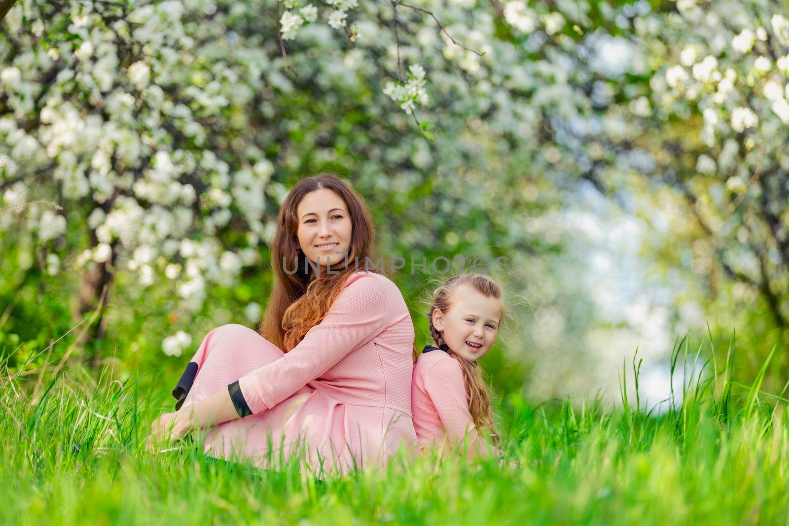 mother and daughter sit in a flowering garden with their backs to each other
