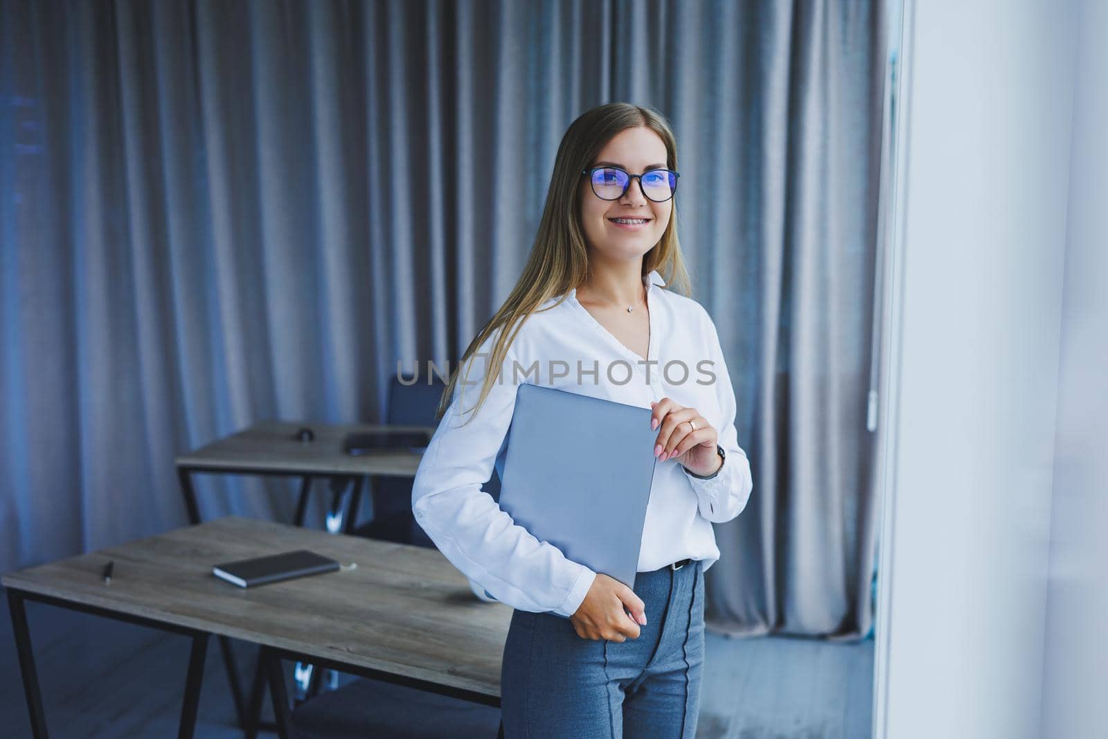 A young smiling business woman is standing in the office by the window with a laptop in her hands. Woman manager in glasses and white shirt by Dmitrytph