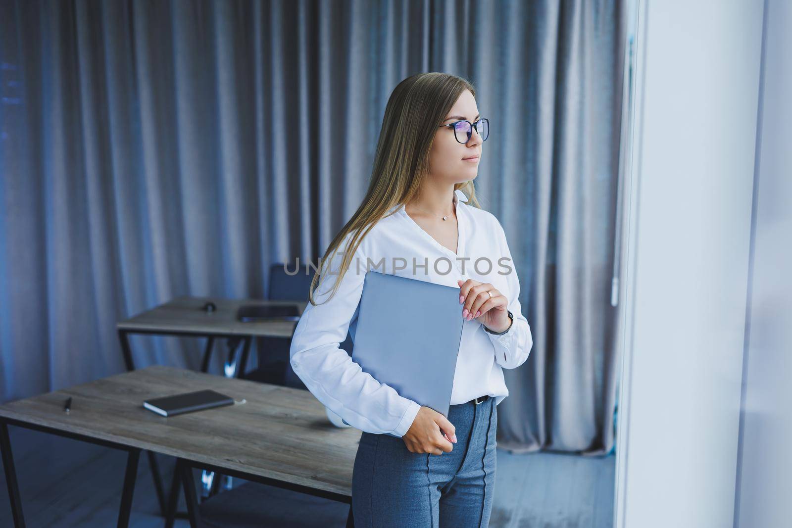 A young smiling business woman is standing in the office by the window with a laptop in her hands. Woman manager in glasses and white shirt