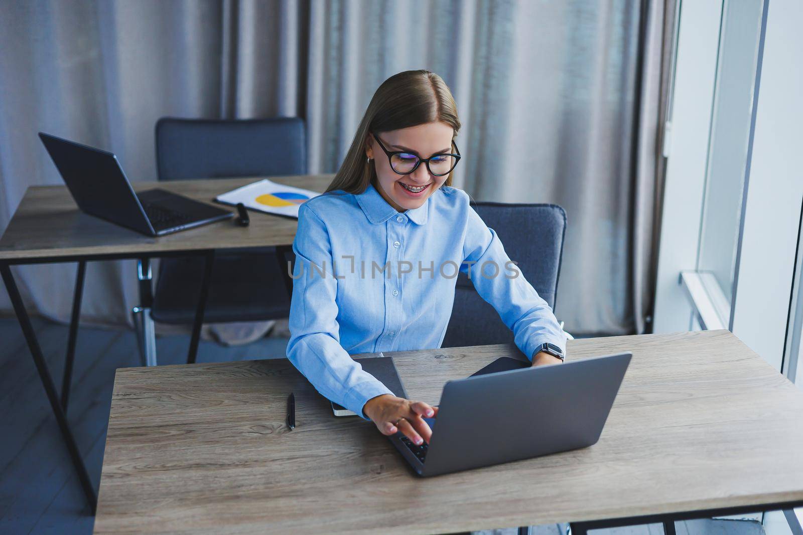 The secretary checks the schedule. Cheerful young beautiful woman in glasses with a smile uses a laptop while sitting at her workplace