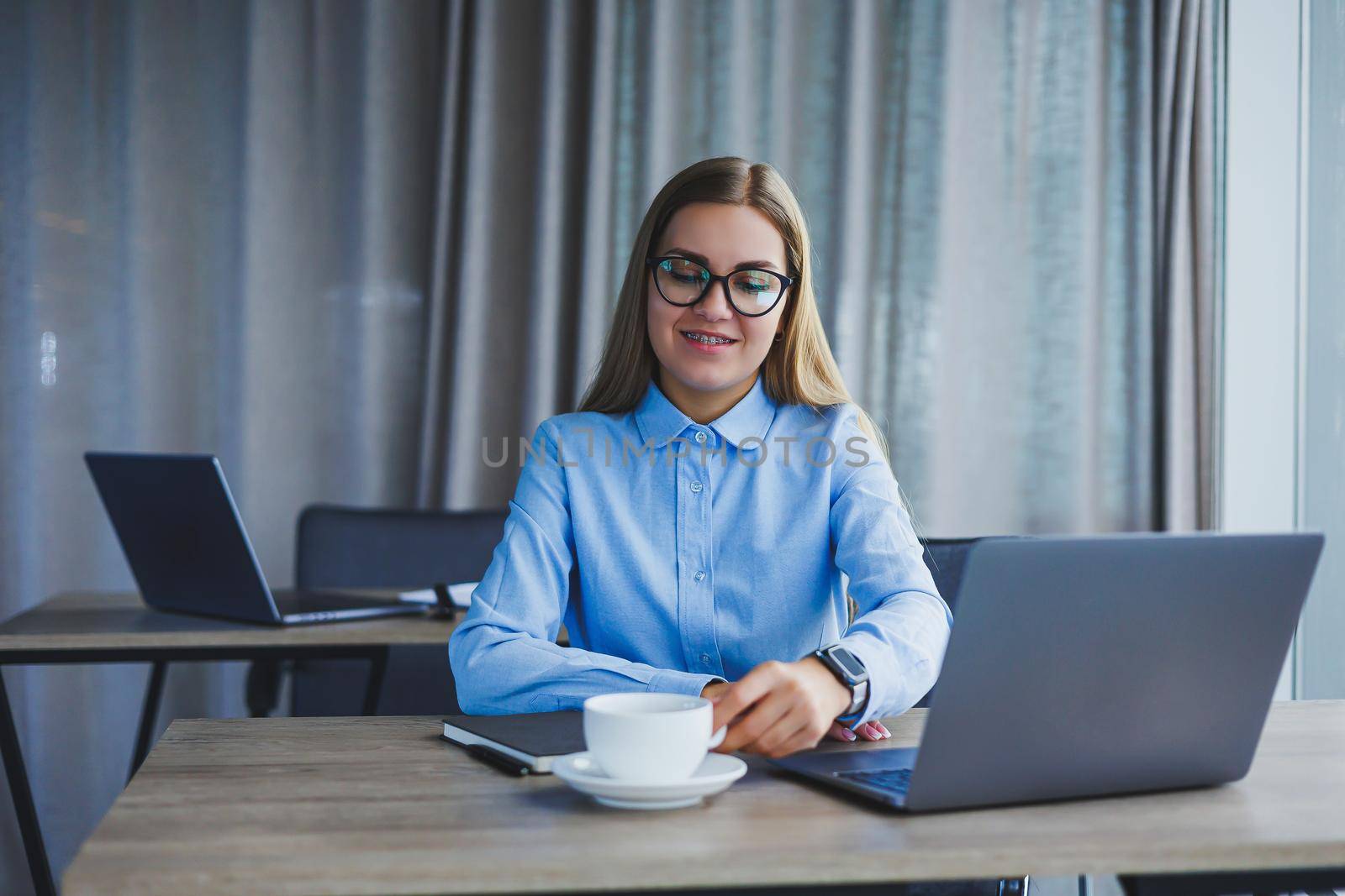 A beautiful business woman in a shirt and glasses sits with a laptop in the office at the table. Woman manager in glasses at workplace in modern office. Working day at the computer