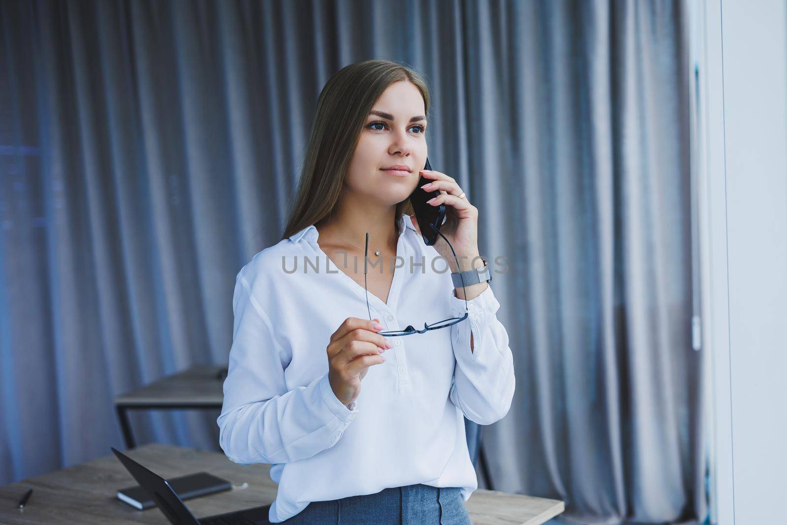 A successful smiling business woman is standing in the office by the window with a phone in her hands. A female manager in glasses and a white shirt is talking on the phone