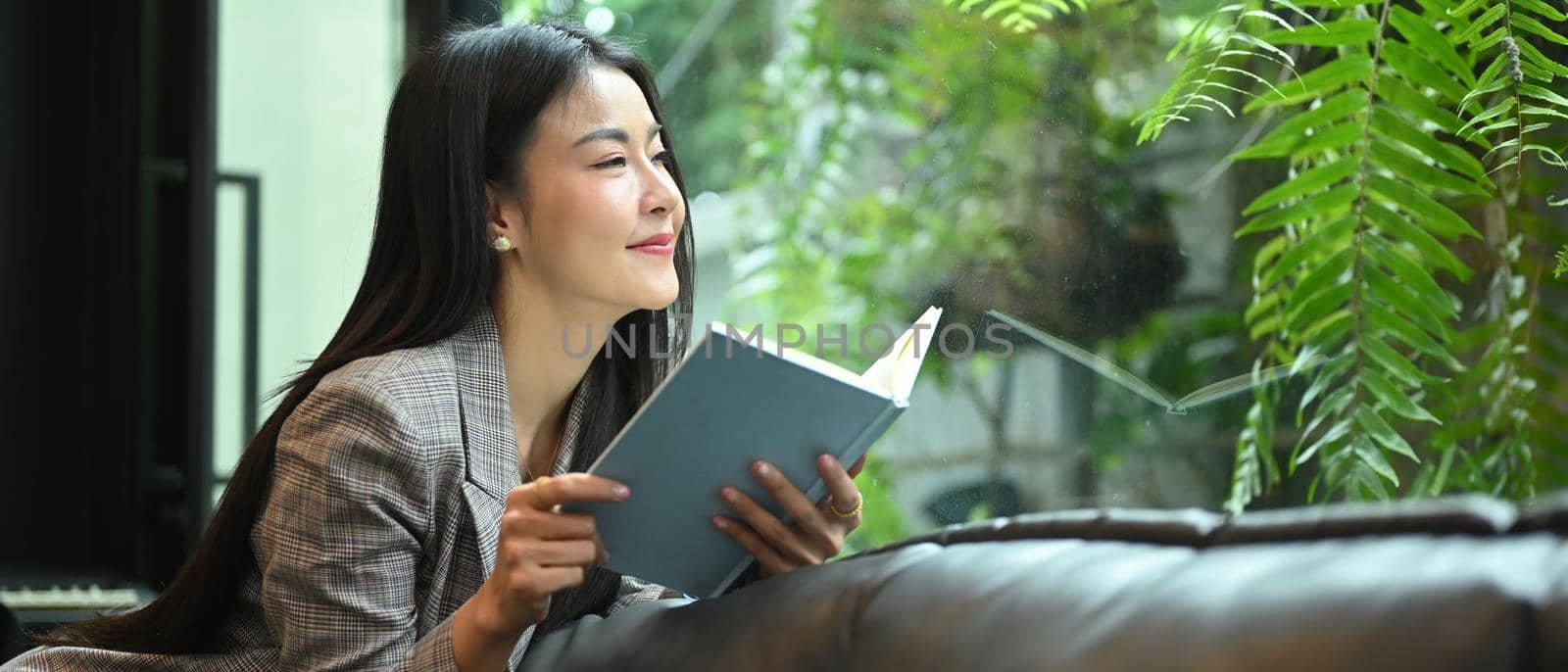 Pleasant millennial business woman resting on couch in bright comfortable office and reading book.