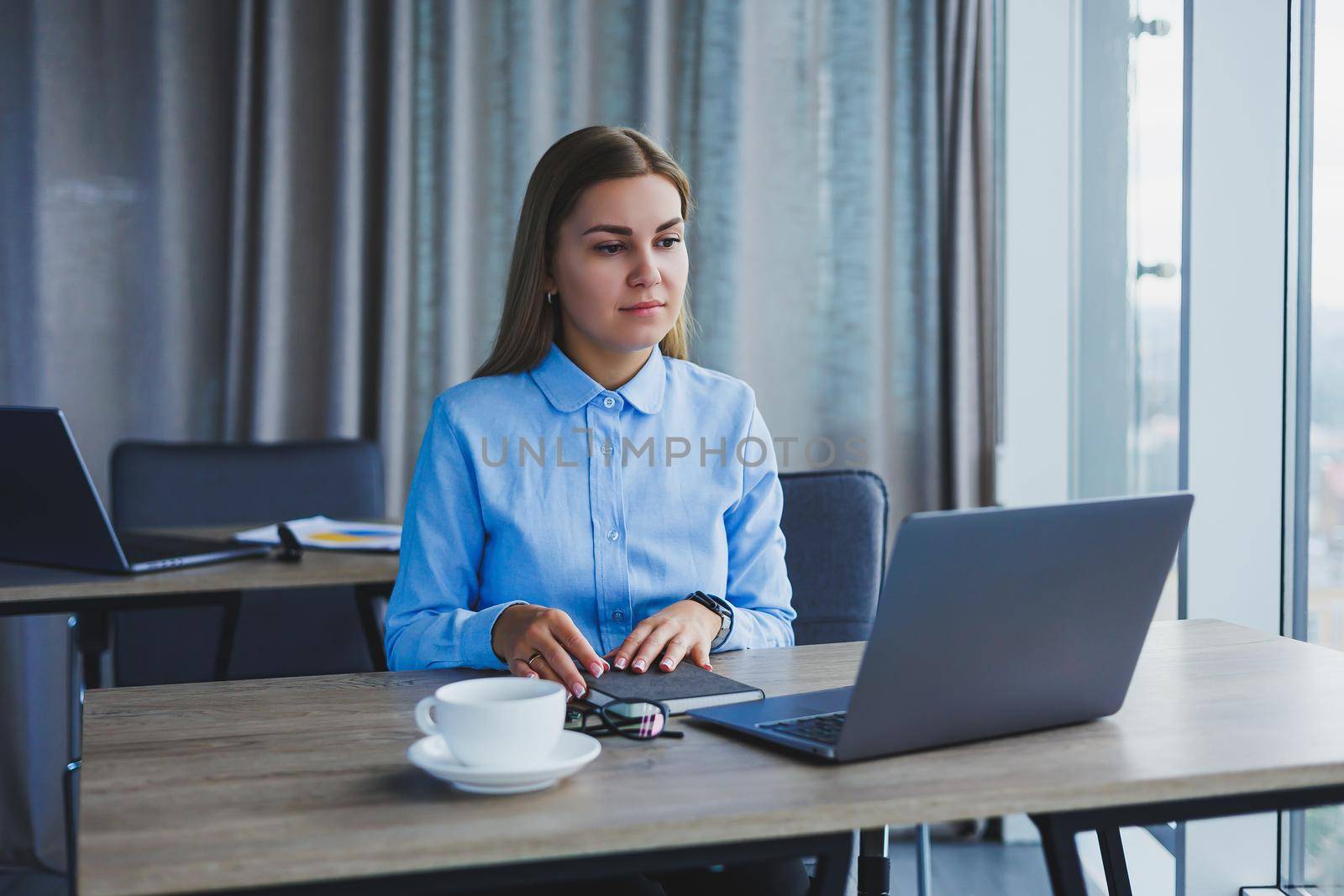 The secretary checks the schedule. Cheerful young beautiful woman in glasses with a smile uses a laptop while sitting at her workplace