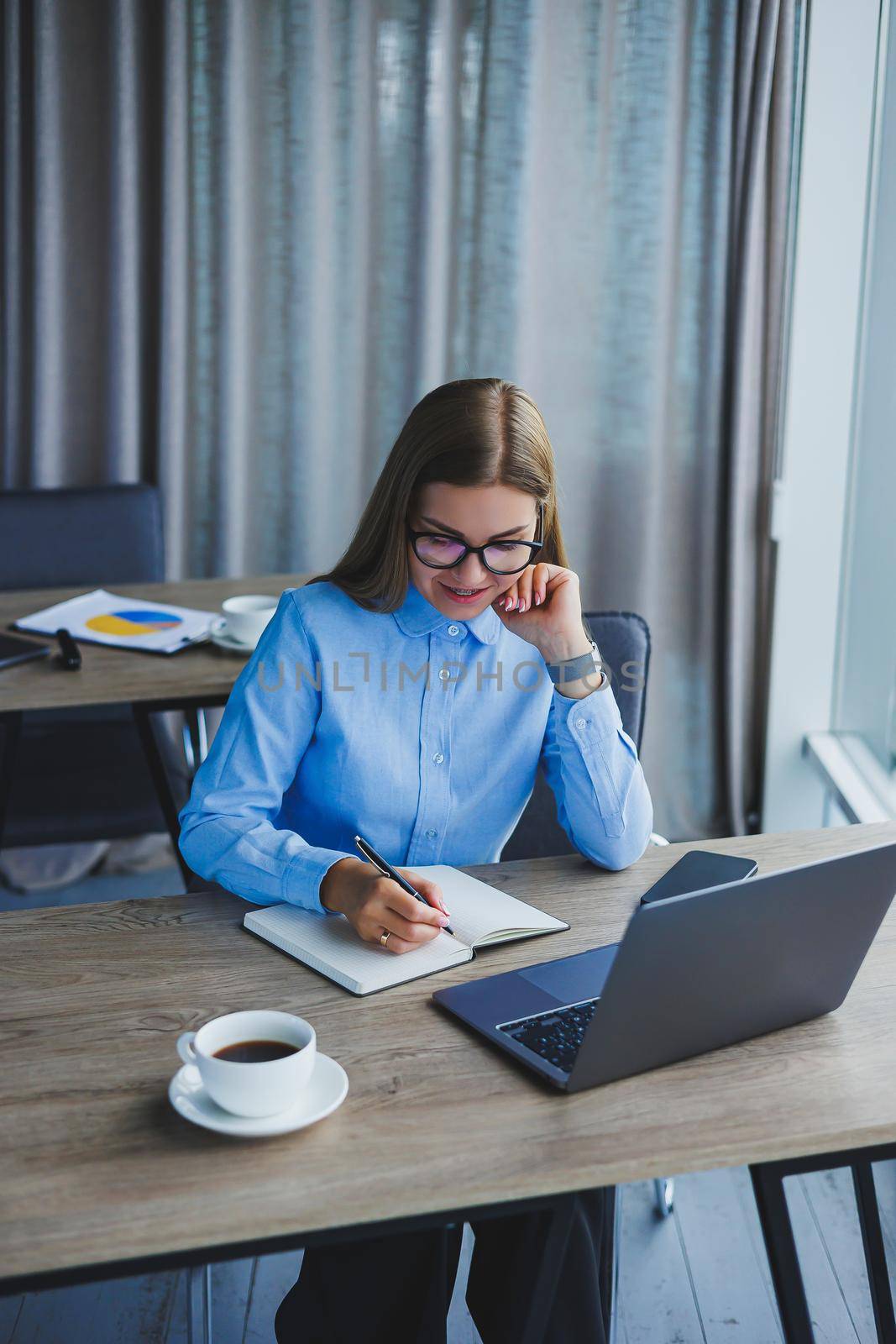 A beautiful business woman in a shirt and glasses sits with a laptop in the office at the table. Woman manager in glasses at workplace in modern office. Working day at the computer