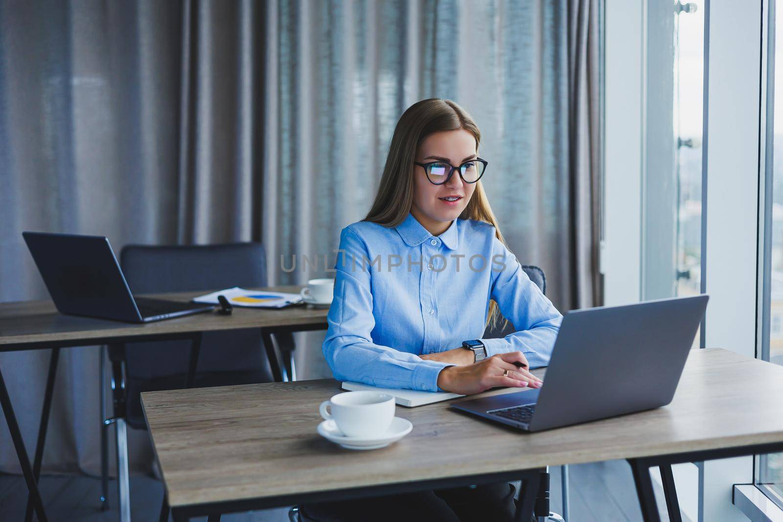 A beautiful business woman in a shirt and glasses sits with a laptop in the office at the table. Woman manager in glasses at workplace in modern office. Working day at the computer
