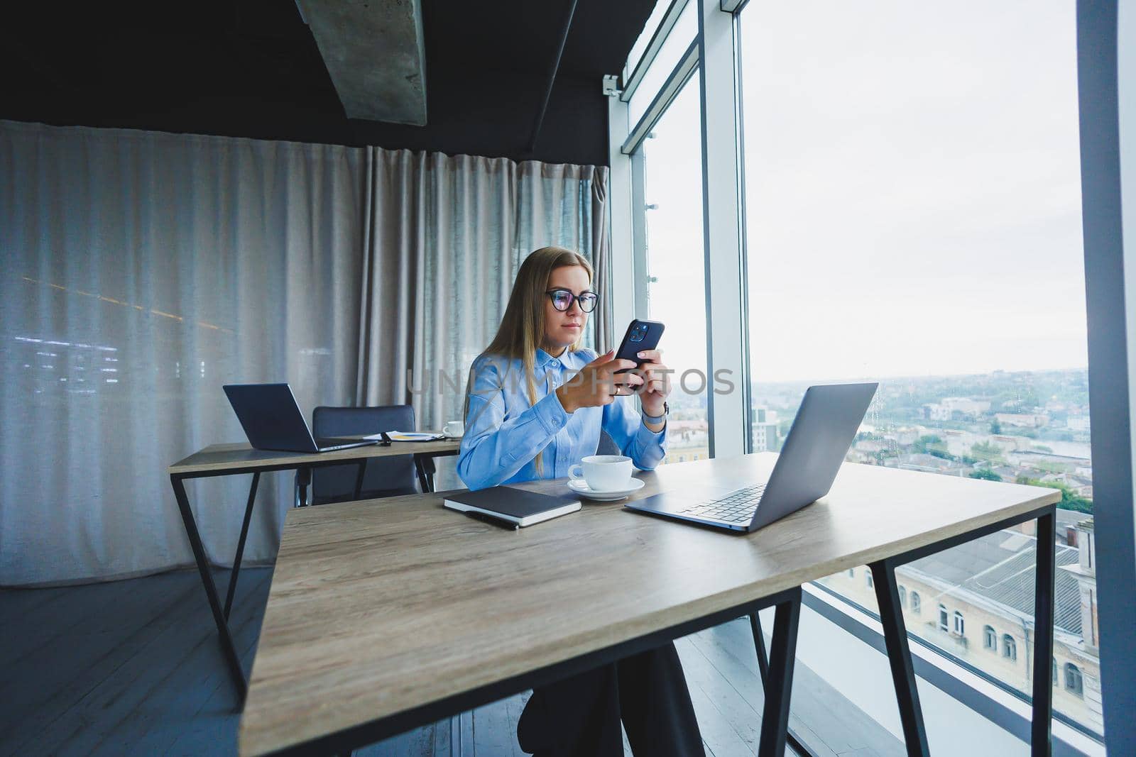 Adult smiling female manager in glasses and formal shirt sits at a desk and looks at a laptop. Modern, spacious, bright office with a large window.