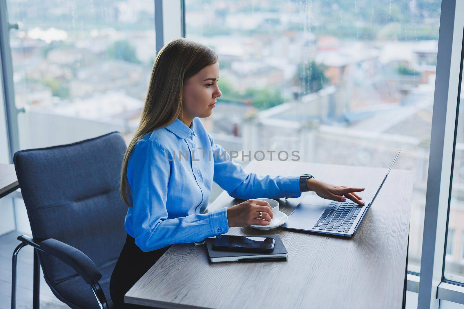 Portrait of cheerful woman in classic glasses smiling in free time in cafe with coffee, positive european woman in blue shirt, desk with laptop, remote work