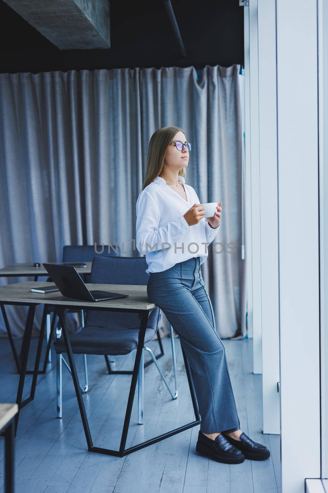 Young smiling business woman standing in office near window with coffee in hands. A female manager in glasses and a white shirt is resting from work