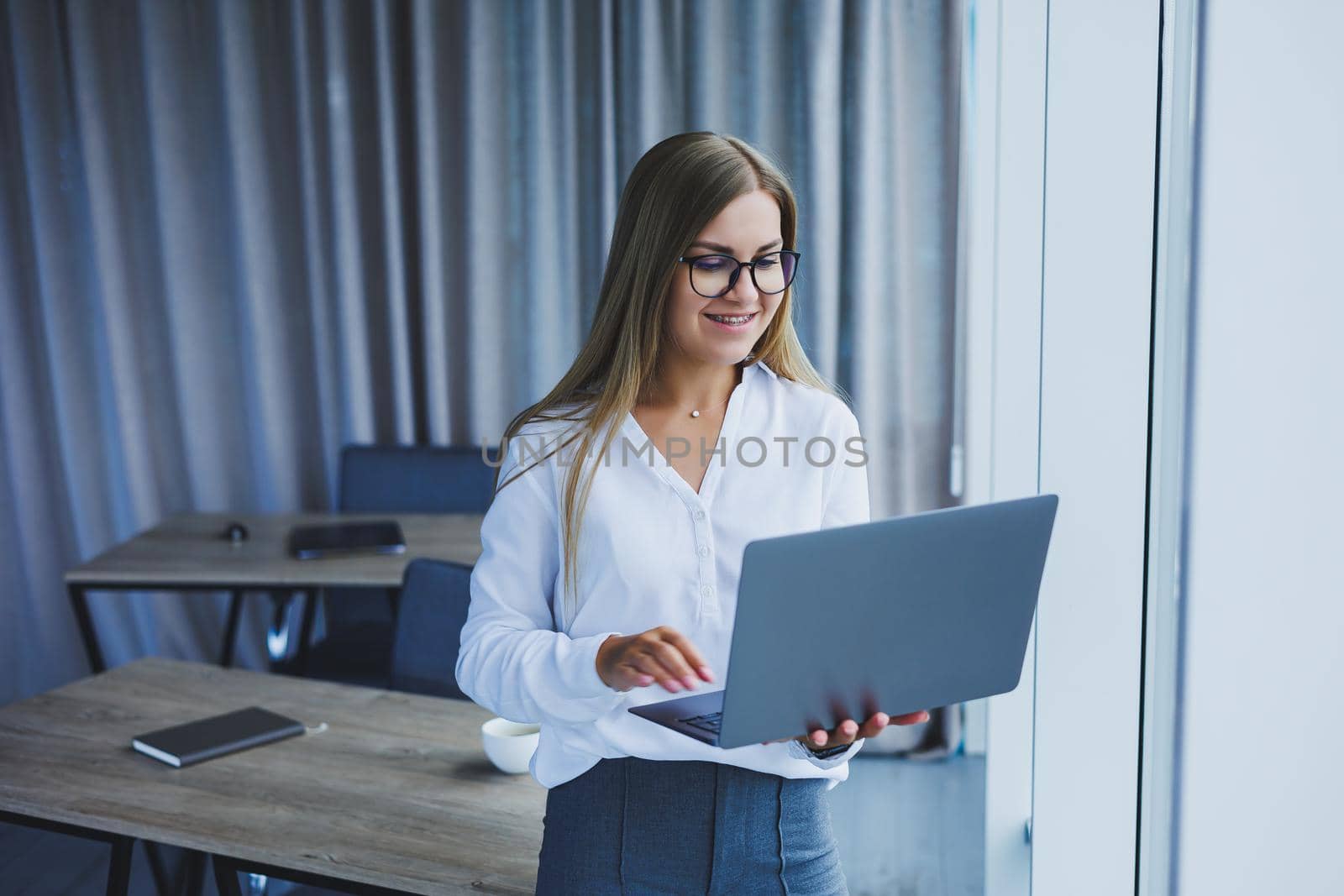 A young smiling business woman is standing in the office by the window with a laptop in her hands. Woman manager in glasses and white shirt by Dmitrytph