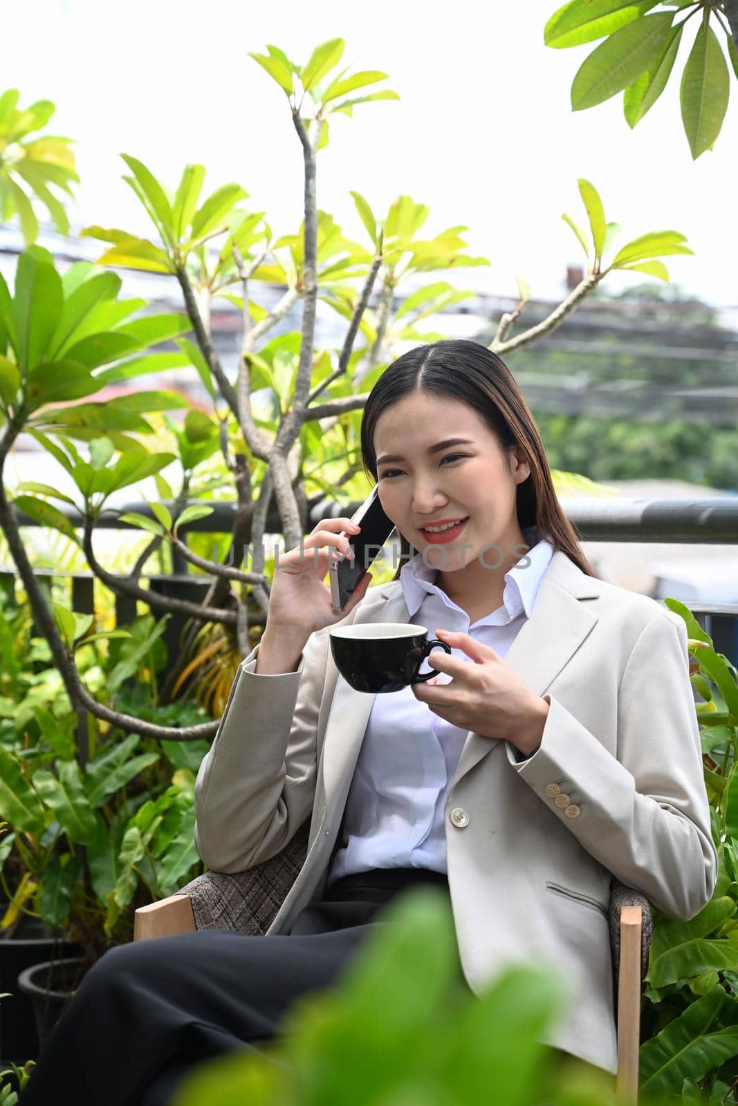 Successful businesswoman having phone conversation while sitting at outdoors cafe surrounded by trees on a beautiful day by prathanchorruangsak