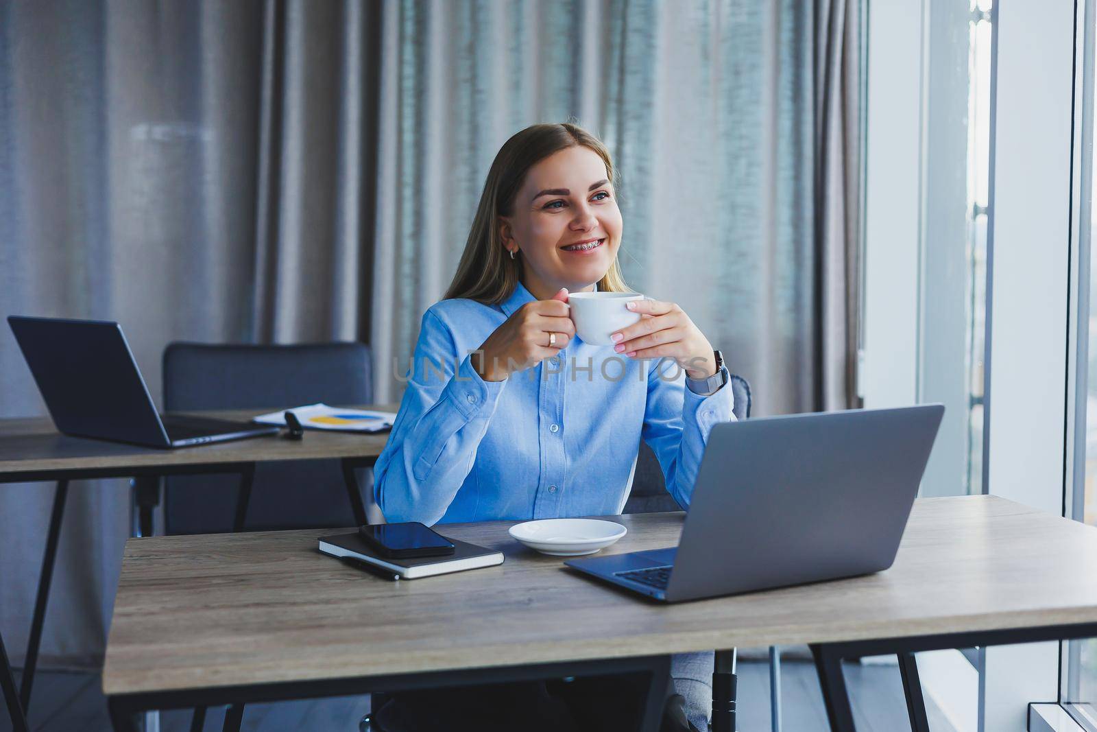 Portrait of cheerful woman in classic glasses smiling in free time in cafe with coffee, positive european woman in blue shirt, desk with laptop, remote work