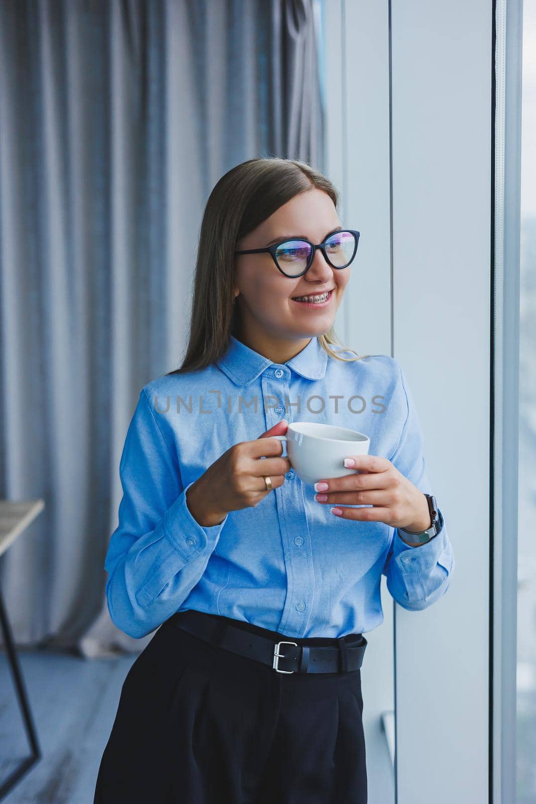 Image of a happy young woman in a classic shirt and glasses smiling and drinking coffee while standing by the window in a modern office with large windows. Remote work