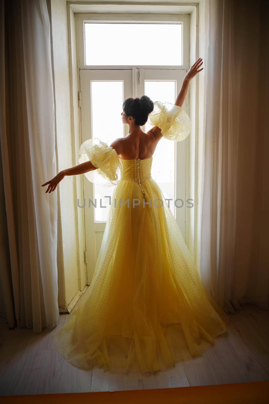 A woman's silhouette in a golden luxurious dress against the background of a window holds a curtain with her hands. Elegant lady in a yellow long silk dress with bare back, back view. by Matiunina