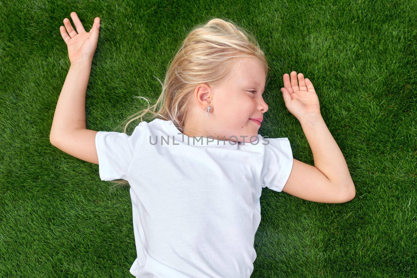 Top view. Mock up for logo, text, design. Blonde long hair child girl lying stretching on green grass. Preschool girl 5-6 years old in white t shirt. Lifestyle Summer vacations Leisure. People